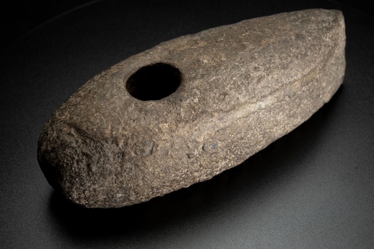 Skafthålsyxa av grovkornig gråbrun bergart. Ett stort och kraftigt exemplar. Plan undersida. Välvda smalsidor, vilka utan begränsningslinjer övergår i ovansidan. Delvis slipad.