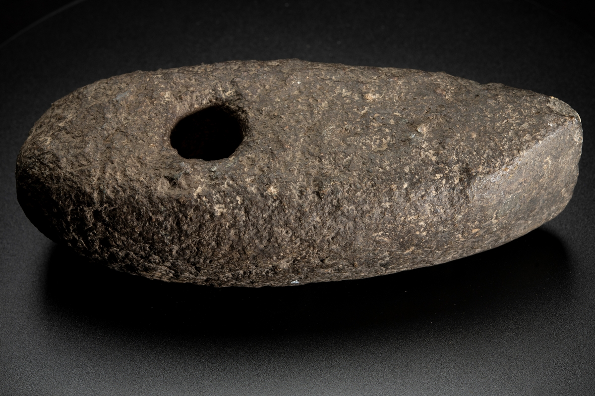 Skafthålsyxa av grovkornig gråbrun bergart. Ett stort och kraftigt exemplar. Plan undersida. Välvda smalsidor, vilka utan begränsningslinjer övergår i ovansidan. Delvis slipad.