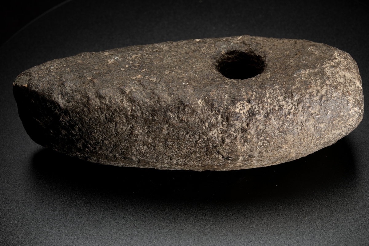 Skafthålsyxa av grovkornig gråbrun bergart. Ett stort och kraftigt exemplar. Plan undersida. Välvda smalsidor, vilka utan begränsningslinjer övergår i ovansidan. Delvis slipad.