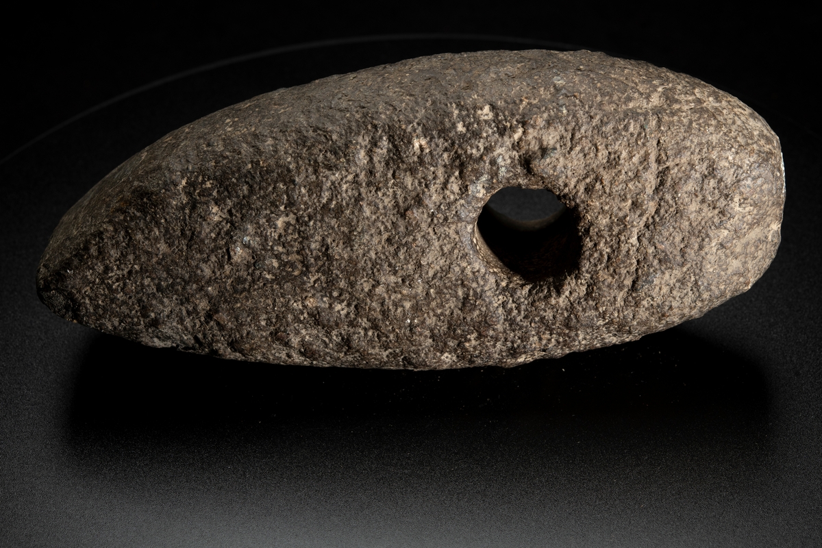 Skafthålsyxa av grovkornig gråbrun bergart. Ett stort och kraftigt exemplar. Plan undersida. Välvda smalsidor, vilka utan begränsningslinjer övergår i ovansidan. Delvis slipad.