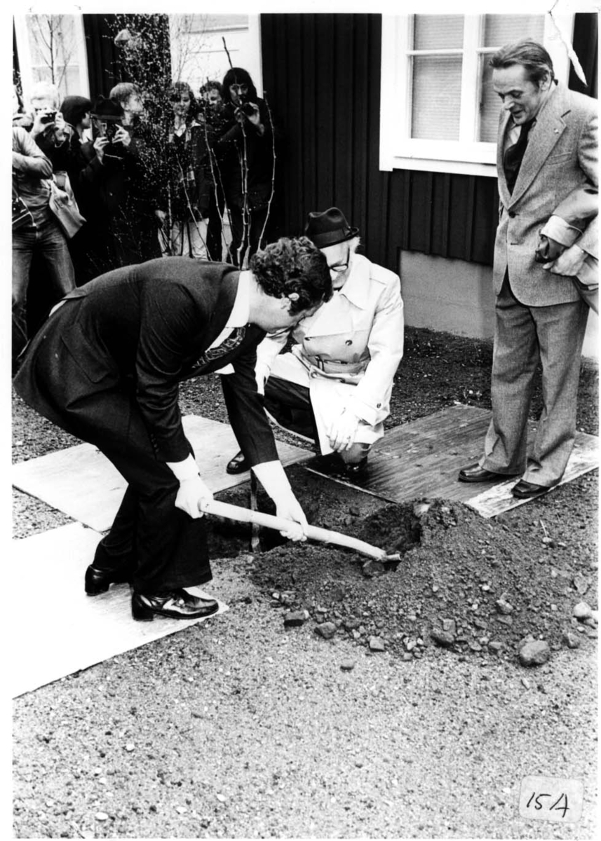 Kung Carl XVI Gustaf gräver en grop utanför Andréemuseet i Gränna. En man i hatt i ljus rock sitter på knä intill och håller en trädplanta som satts i gropen. En annan man står till höger. Folk i bakgrunden som fotograferar.