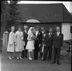 Ett bröllopssällskap står uppställda för fotografering. I mitten står bruden och brudgummen. I bakgrunden syns en kyrka med spånfasad.