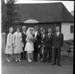 Ett bröllopssällskap står uppställda för fotografering. I mitten står bruden och brudgummen. I bakgrunden syns en kyrka med spånfasad.