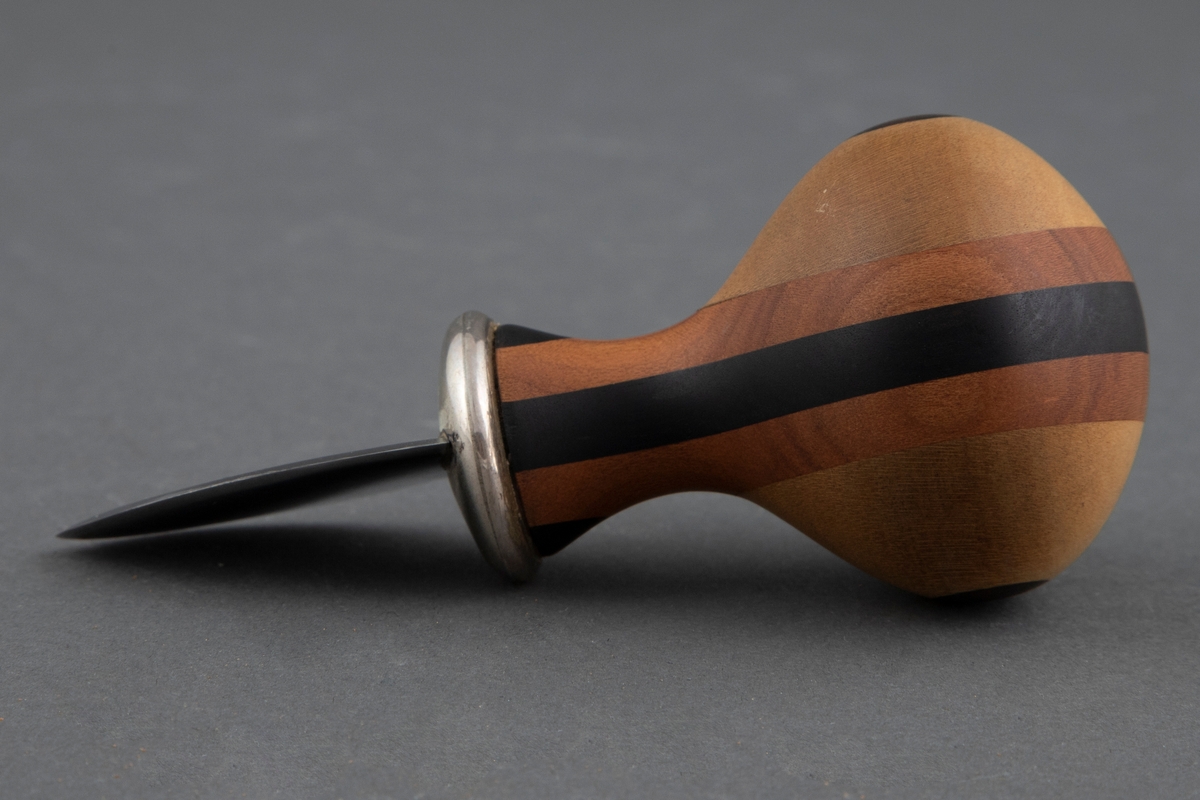 Kniv med kort, fyldig skaft og lite, skjoldformet blad uten skjæreflate. Skaftet er formet av laminert tre av ulike sorter og farger, slik at det danner et regelmessig geometrisk mønster. Bladet er i smidd stål og overgangen mellom skaft og blad er i sølv.