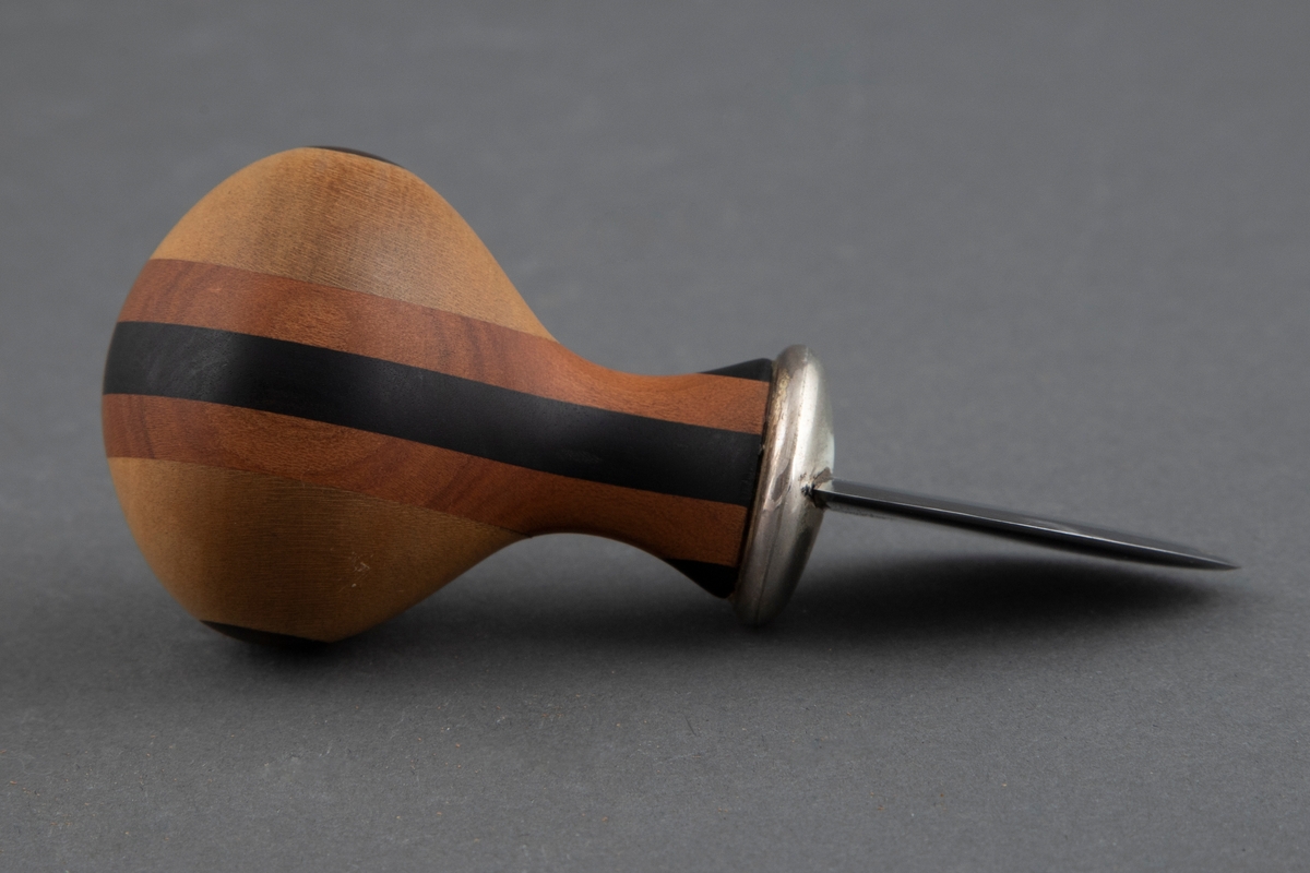 Kniv med kort, fyldig skaft og lite, skjoldformet blad uten skjæreflate. Skaftet er formet av laminert tre av ulike sorter og farger, slik at det danner et regelmessig geometrisk mønster. Bladet er i smidd stål og overgangen mellom skaft og blad er i sølv.