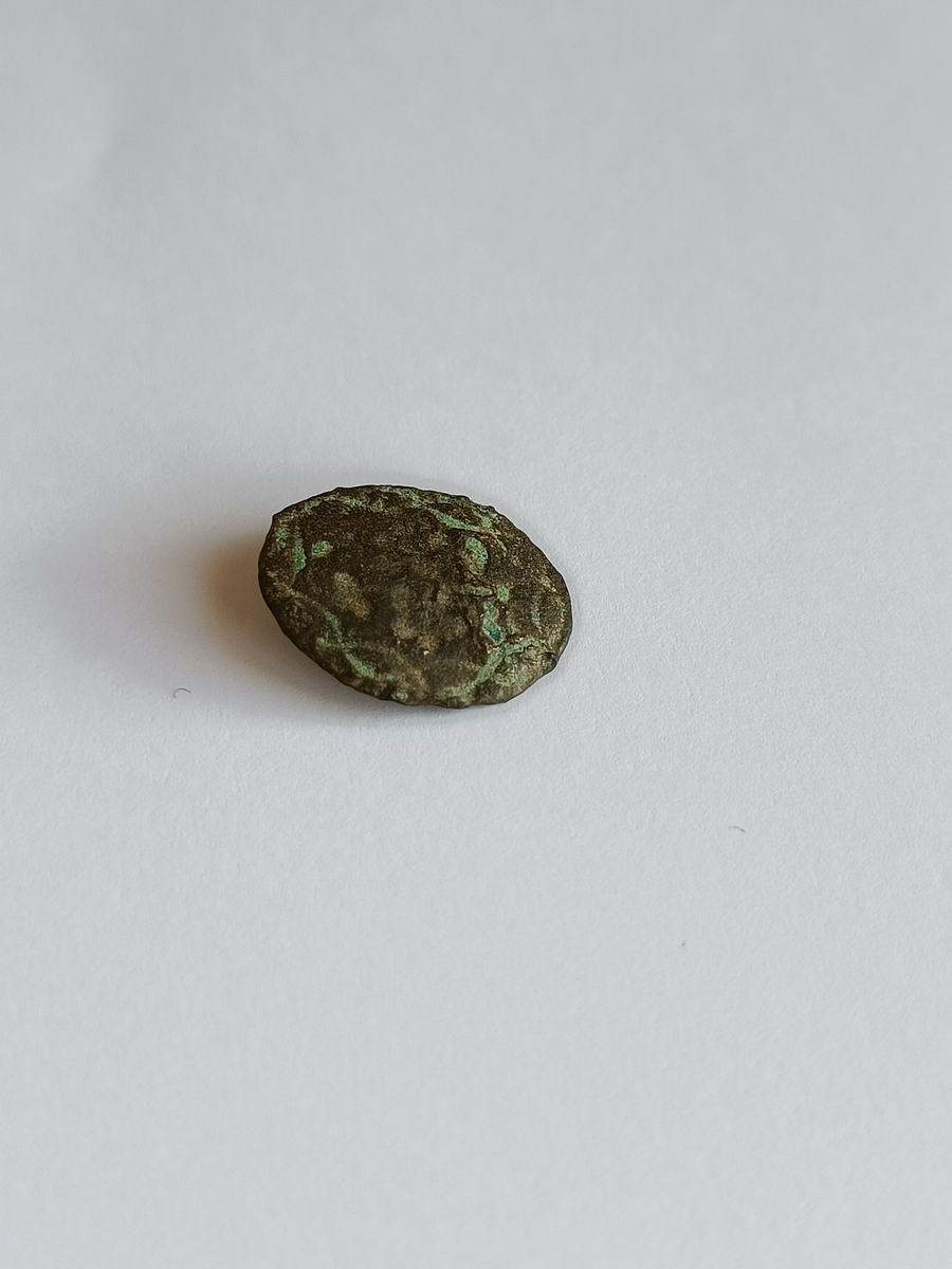 Oval og flat knapp med rester av strekdekor i et flettemønster langs kantene, og med en fin hempe på baksiden.