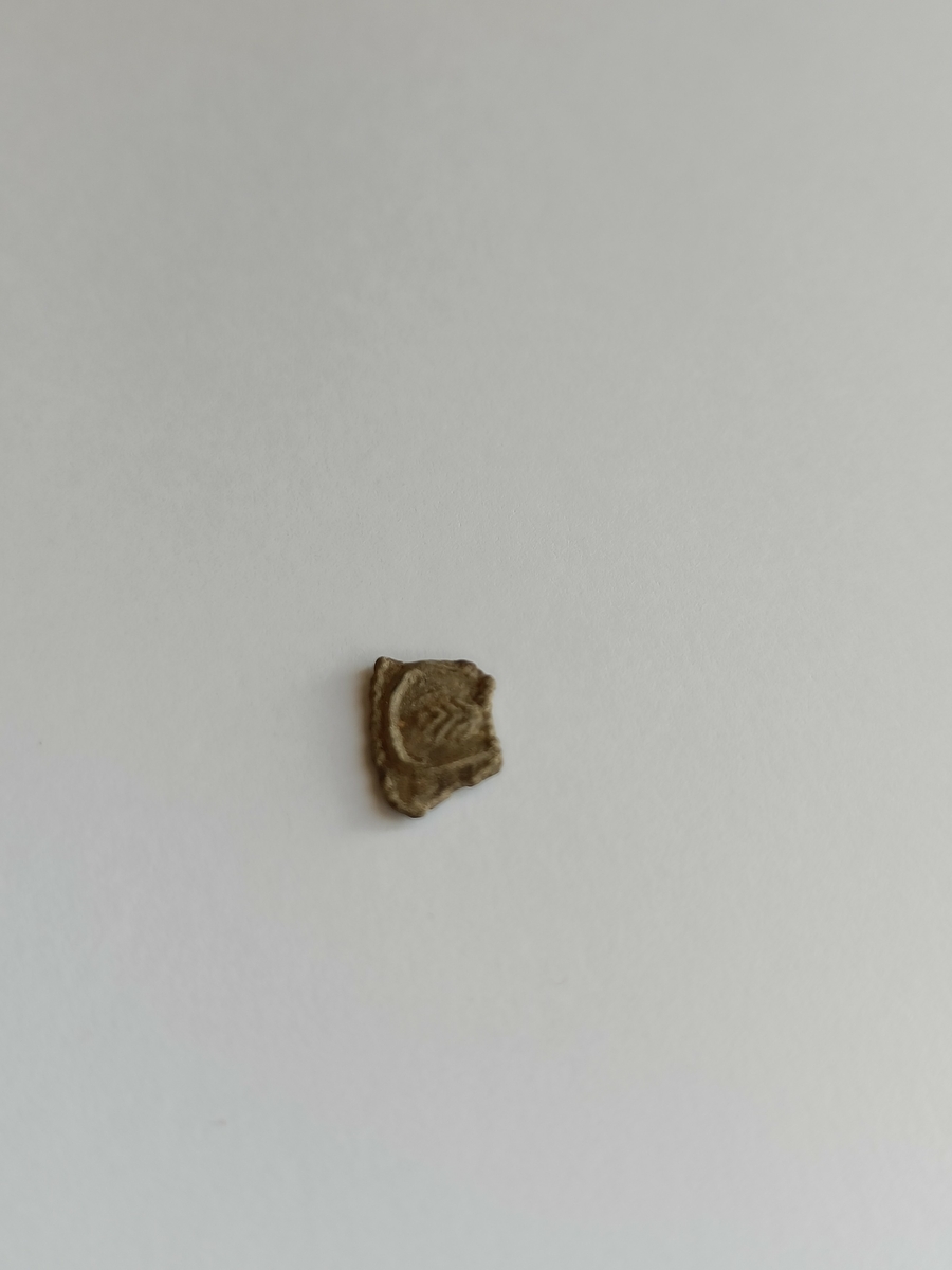 Liten og tilnærmet kvadratisk del med bruddflater i to ender og krumming av de to andre sidene som også har en langsgående perlerad.  Beslagsbiten dekkes av et motiv av et trekantet bladomkranset av en linje. Motivet er opphøyd.