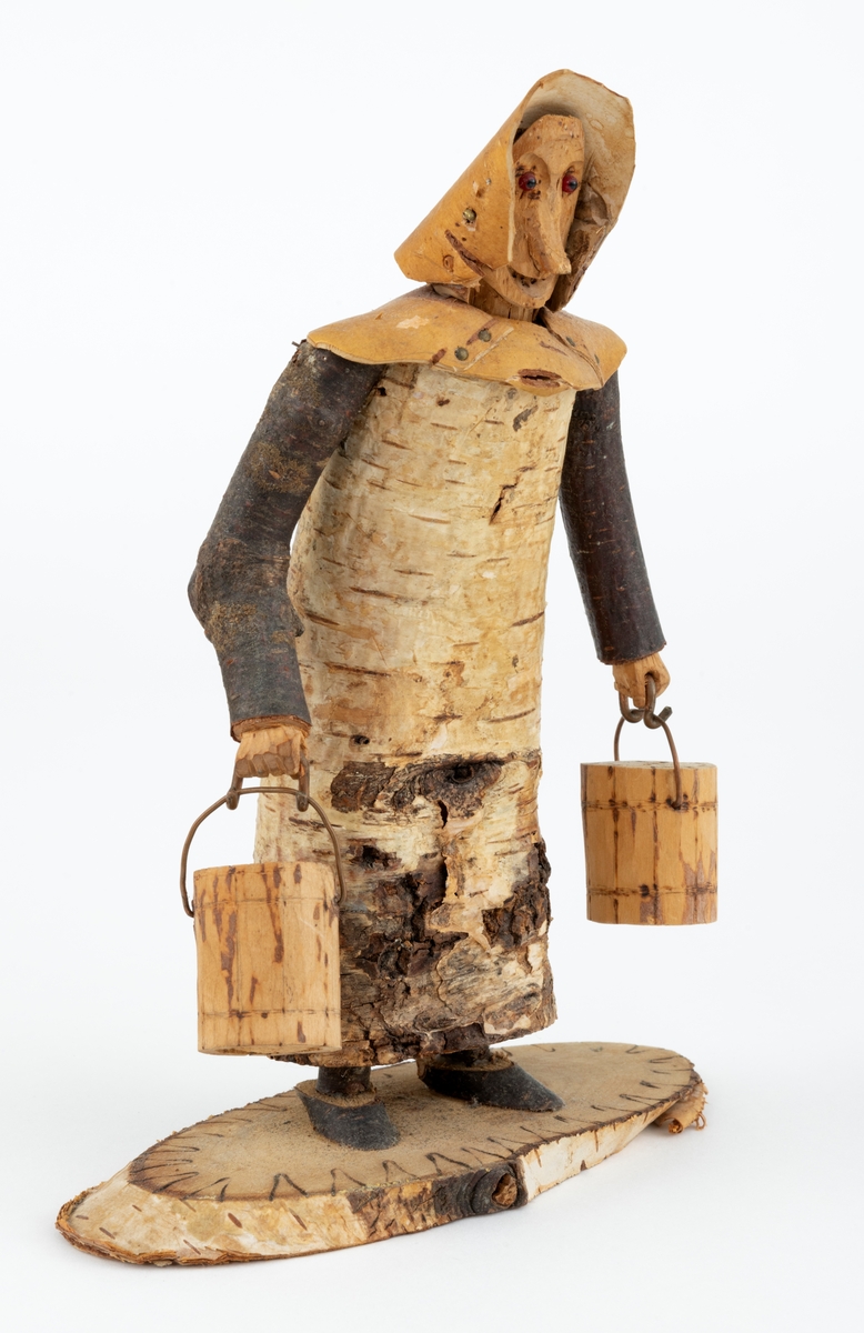 Trefigur spikket av tre, med påmontert never. Figuren er festet til en treplate.
Figuren er en etterligning etter moren til Eldbjørg Kordal, Ellen Pauline (f. Einarsrud) Kordal. Kona bærer ei vannbøtte i hver hand. Trefiguren var en gave fra Eldbjørg og hennes far jula 1967.