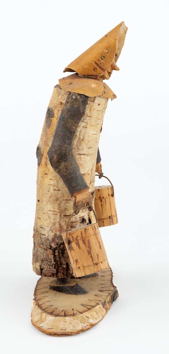 Trefigur spikket av tre, med påmontert never. Figuren er festet til en treplate.
Figuren er en etterligning etter moren til Eldbjørg Kordal, Ellen Pauline (f. Einarsrud) Kordal. Kona bærer ei vannbøtte i hver hand. Trefiguren var en gave fra Eldbjørg og hennes far jula 1967.