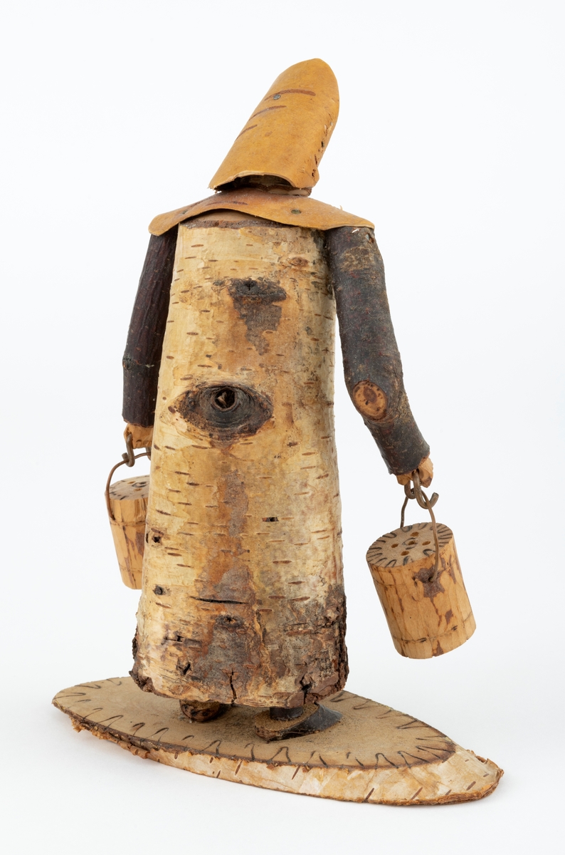 Trefigur spikket av tre, med påmontert never. Figuren er festet til en treplate.
Figuren er en etterligning etter moren til Eldbjørg Kordal, Ellen Pauline (f. Einarsrud) Kordal. Kona bærer ei vannbøtte i hver hand. Trefiguren var en gave fra Eldbjørg og hennes far jula 1967.