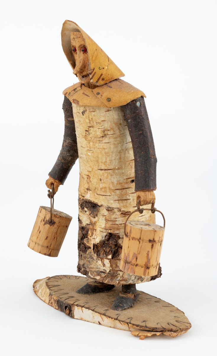 Trefigur spikket av tre, med påmontert never. Figuren er festet til en treplate.
Figuren er en etterligning etter moren til Eldbjørg Kordal, Ellen Pauline (f. Einarsrud) Kordal. Kona bærer ei vannbøtte i hver hand. Trefiguren var en gave fra Eldbjørg og hennes far jula 1967.