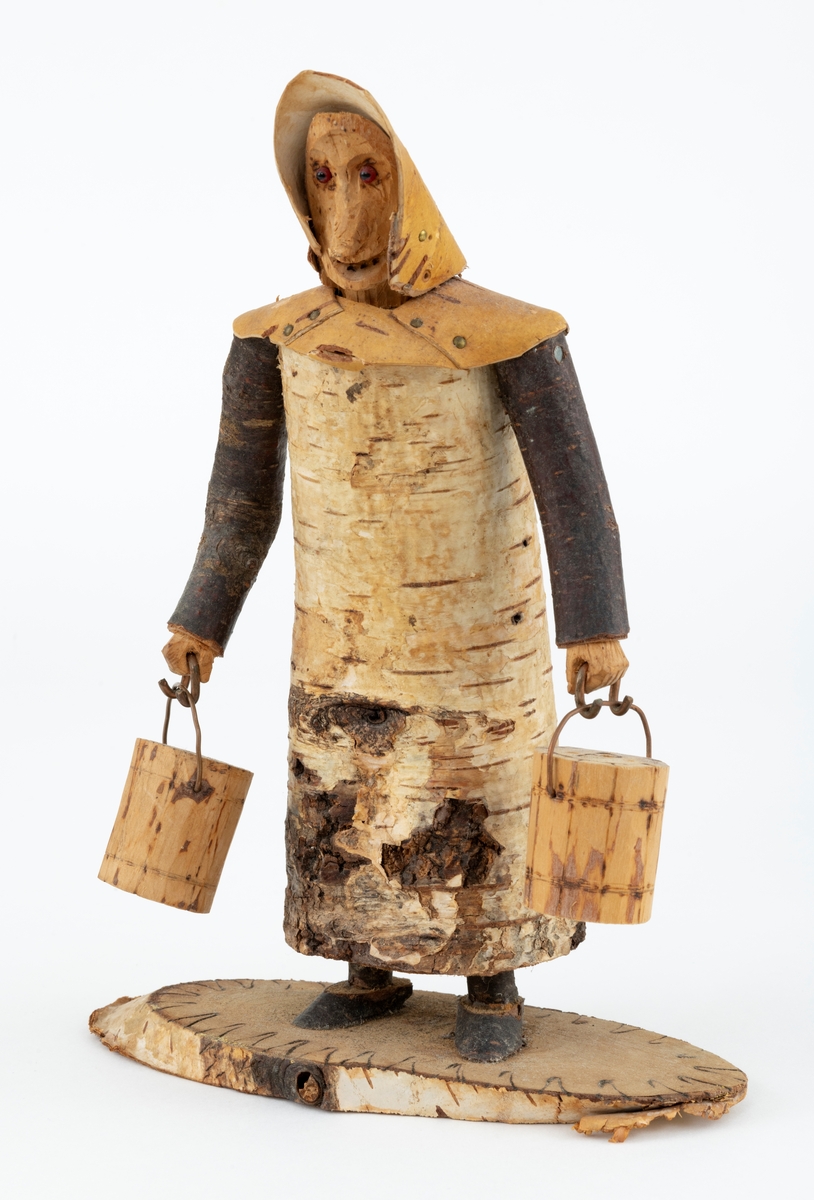 Trefigur spikket av tre, med påmontert never. Figuren er festet til en treplate.
Figuren er en etterligning etter moren til Eldbjørg Kordal, Ellen Pauline (f. Einarsrud) Kordal. Kona bærer ei vannbøtte i hver hand. Trefiguren var en gave fra Eldbjørg og hennes far jula 1967.