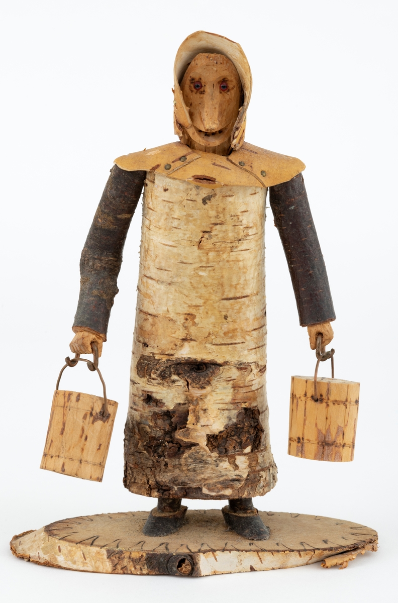 Trefigur spikket av tre, med påmontert never. Figuren er festet til en treplate.
Figuren er en etterligning etter moren til Eldbjørg Kordal, Ellen Pauline (f. Einarsrud) Kordal. Kona bærer ei vannbøtte i hver hand. Trefiguren var en gave fra Eldbjørg og hennes far jula 1967.