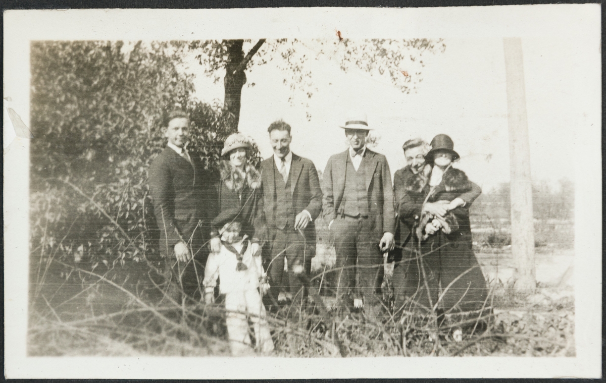 En gruppe menn, kvinner og et barn smiler til fotografen i en park. Fra venstre er Jakob Lura, mrs. Ragnhild Baug med gutten Billy, Bjarne Fixdal, mr. Baug, Karl Bårdsen og ukjent.