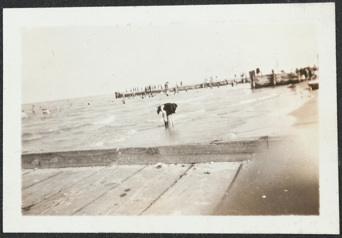 Kvinne på stranden, Calvary beach, 1924. Pir eller kai i forgrunnen og flere i bakgrunnen Man skimter mange mennesker på stranden og badende i havet.