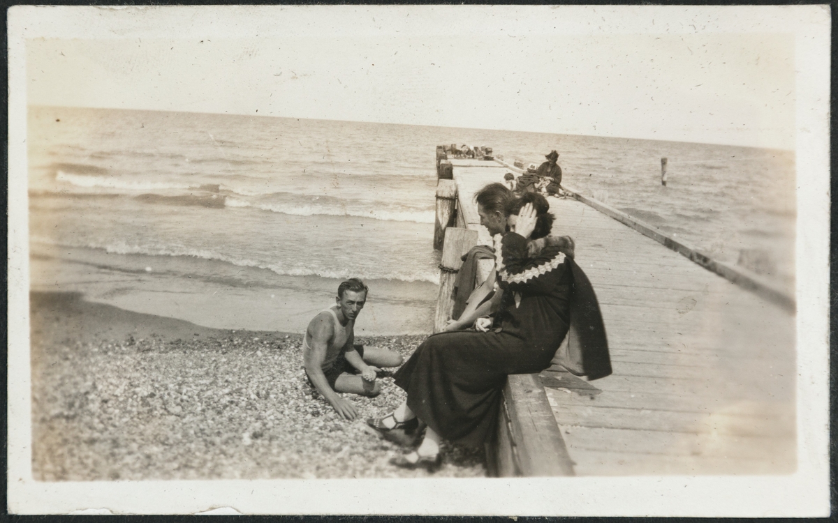 To menn og en kvinne på stranden, Calvary beach, 1924. Pir eller kai i forgrunnen og flere i bakgrunnen Man skimter mange mennesker på stranden og badende i havet. Kalle (Karl Bårdsen) sitter på stranda.