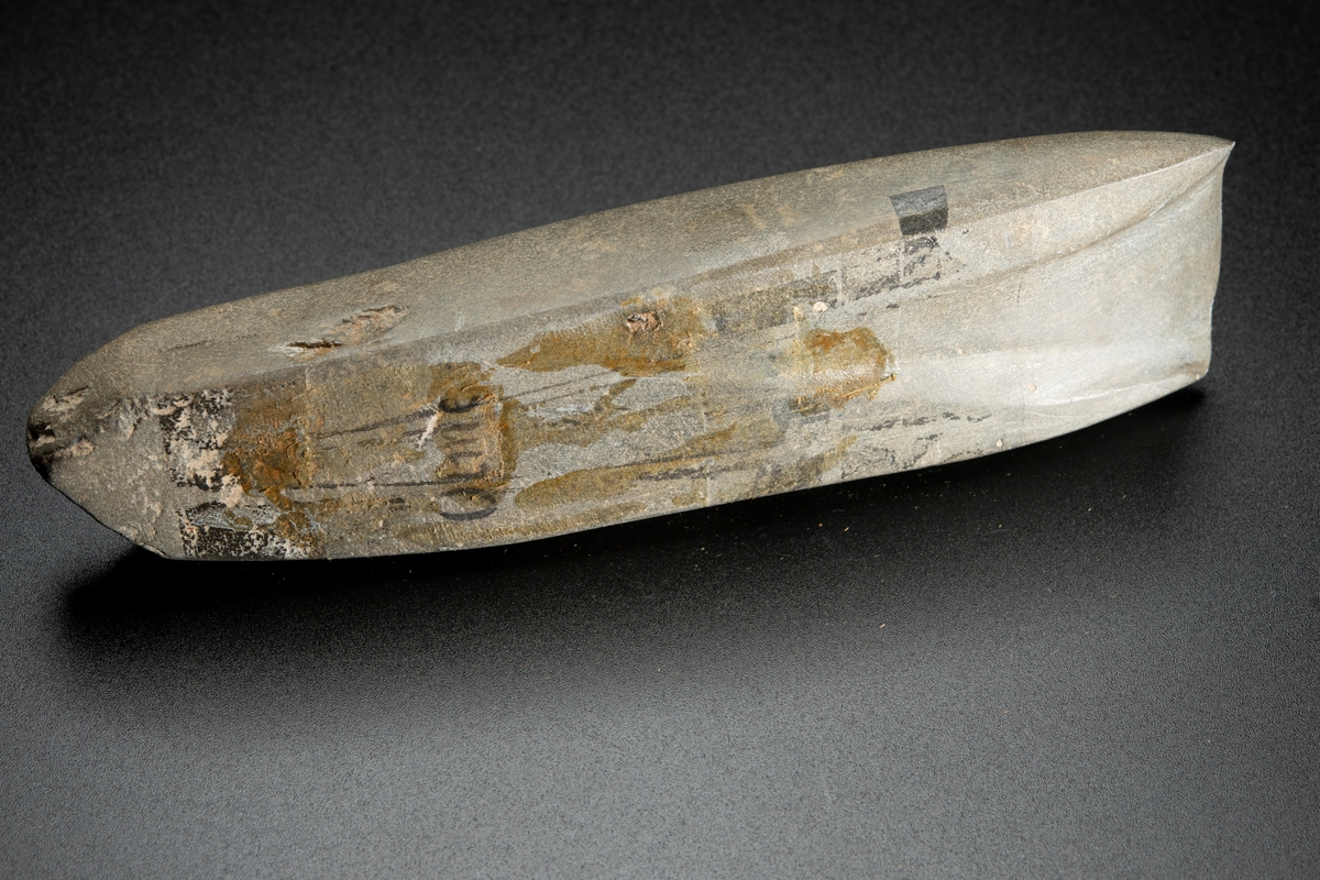 I det närmaste jämnbred. Fyrsidig. Kraftig hålslipning. Helt slipad. Av finkornig bergart.