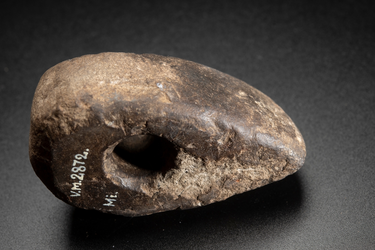 Av finkornig bergart. Delvis slipad. Oregelbundet nackparti. Plan ovan- och undersida, i det närmaste raka sidor.