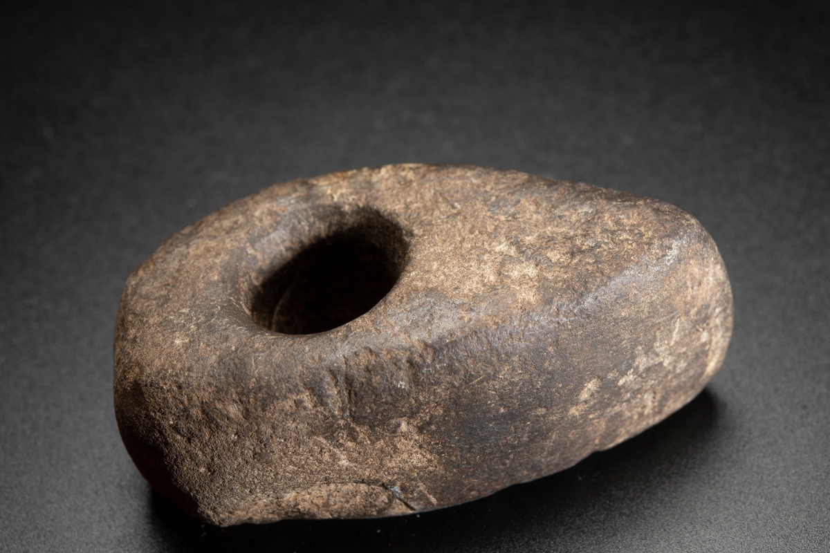 Av finkornig bergart. Delvis slipad. Oregelbundet nackparti. Plan ovan- och undersida, i det närmaste raka sidor.