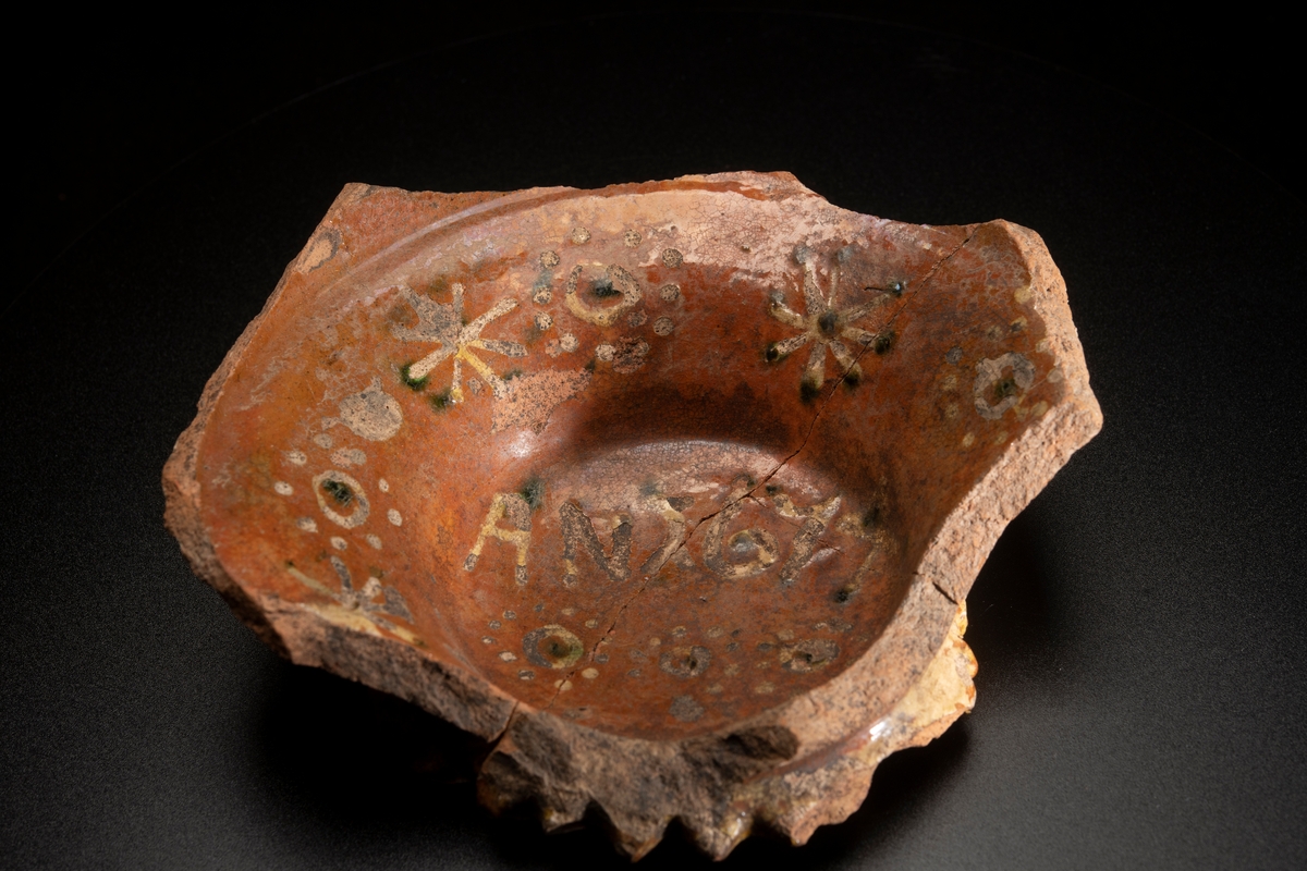 Lerskål, med delar av sidorna borta. In- och utvändig glacyr tegelfärgad med dekor i gult och grönt. Utsidans dekor bestående av ränder i gult , insidans av ringar och stjärnor i gult och grönt. I botten står " AN 1674 " i samma färger. Botten i ytterkanten prydd med uddar och utanpå konvex vilket tyder på att skålen varit försedd med fötter, efter vilka även märken finnes. Tre hopsatta skärvor.