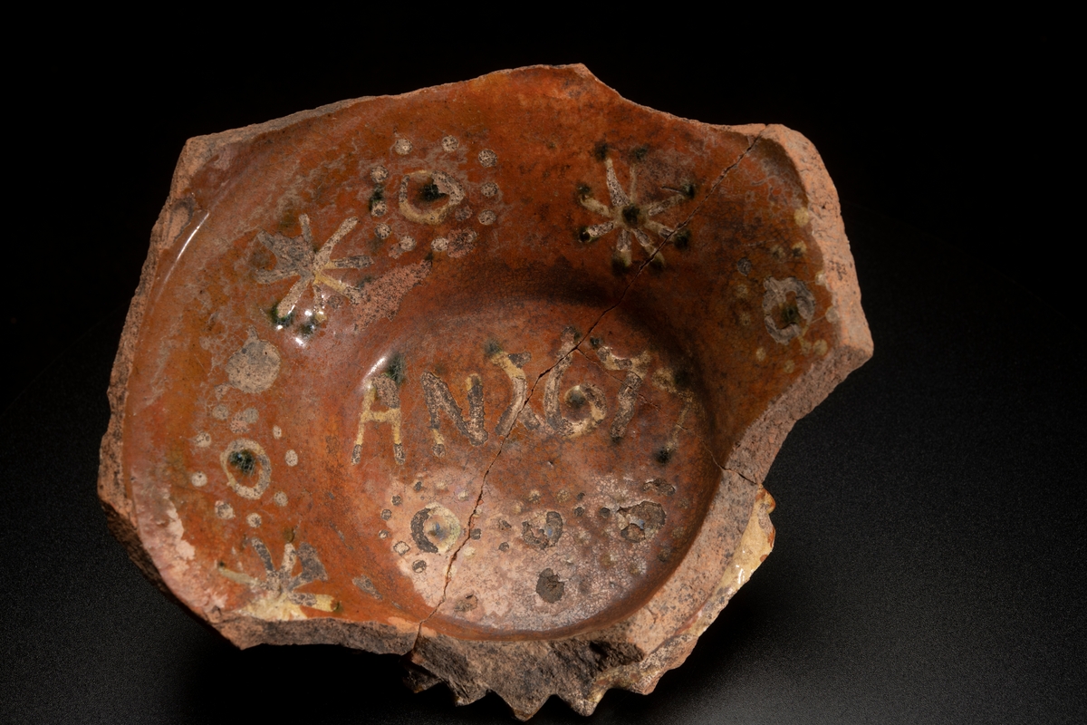 Lerskål, med delar av sidorna borta. In- och utvändig glacyr tegelfärgad med dekor i gult och grönt. Utsidans dekor bestående av ränder i gult , insidans av ringar och stjärnor i gult och grönt. I botten står " AN 1674 " i samma färger. Botten i ytterkanten prydd med uddar och utanpå konvex vilket tyder på att skålen varit försedd med fötter, efter vilka även märken finnes. Tre hopsatta skärvor.