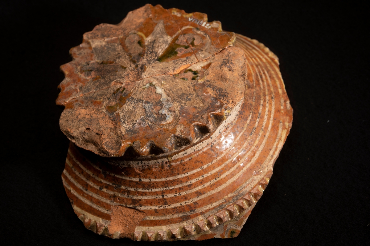 Lerskål, med delar av sidorna borta. In- och utvändig glacyr tegelfärgad med dekor i gult och grönt. Utsidans dekor bestående av ränder i gult , insidans av ringar och stjärnor i gult och grönt. I botten står " AN 1674 " i samma färger. Botten i ytterkanten prydd med uddar och utanpå konvex vilket tyder på att skålen varit försedd med fötter, efter vilka även märken finnes. Tre hopsatta skärvor.