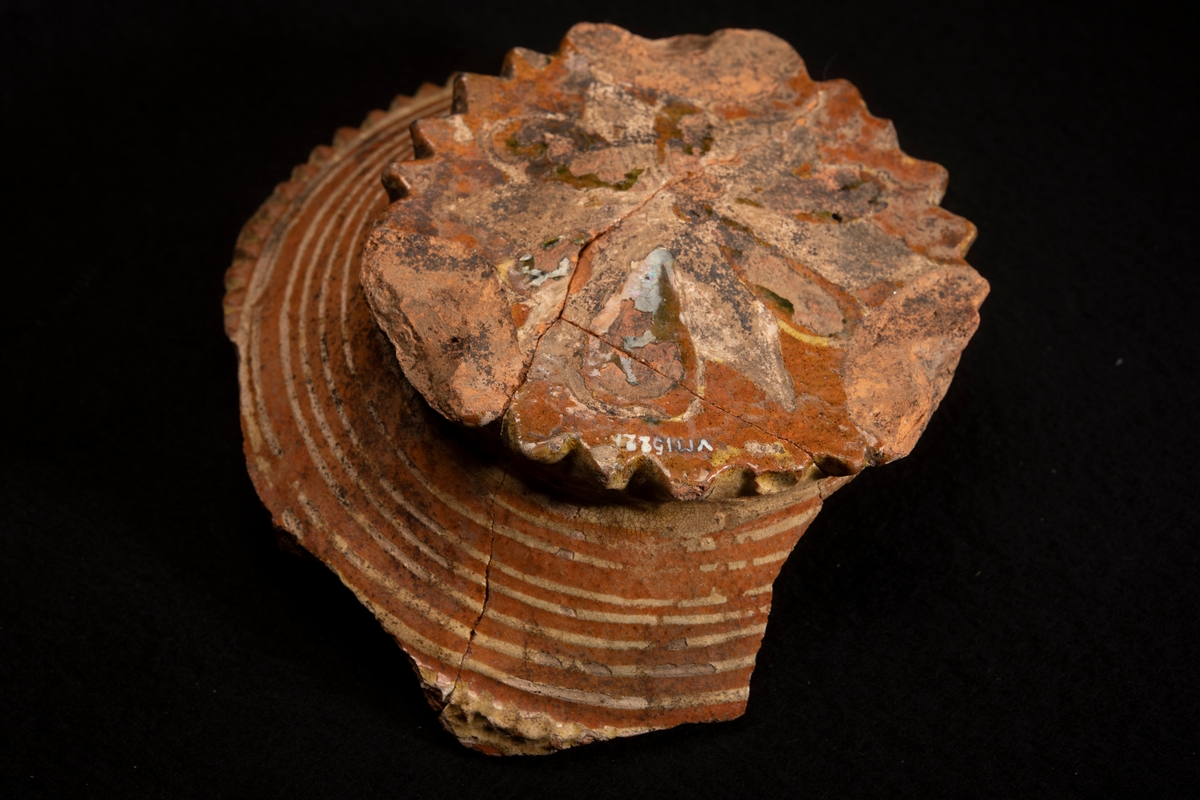 Lerskål, med delar av sidorna borta. In- och utvändig glacyr tegelfärgad med dekor i gult och grönt. Utsidans dekor bestående av ränder i gult , insidans av ringar och stjärnor i gult och grönt. I botten står " AN 1674 " i samma färger. Botten i ytterkanten prydd med uddar och utanpå konvex vilket tyder på att skålen varit försedd med fötter, efter vilka även märken finnes. Tre hopsatta skärvor.