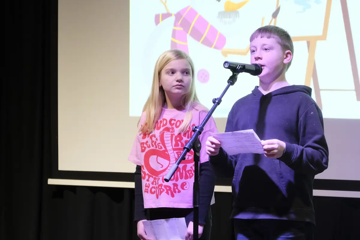 En gutt og ei jente fra femte klasse ved Eidsvoll Verk skole står ved en mikrofon på et stativ og holder appell. Det skjer i auditoriet i Wergelands hus.