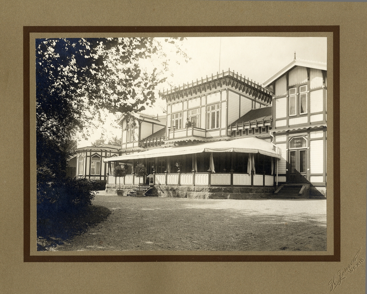 Järnvägsrestaurangen i Växjö en varm sommardag i början av 1900-talet. 
En man (möjl. källarmästaren) syns på restaurangens balkong.