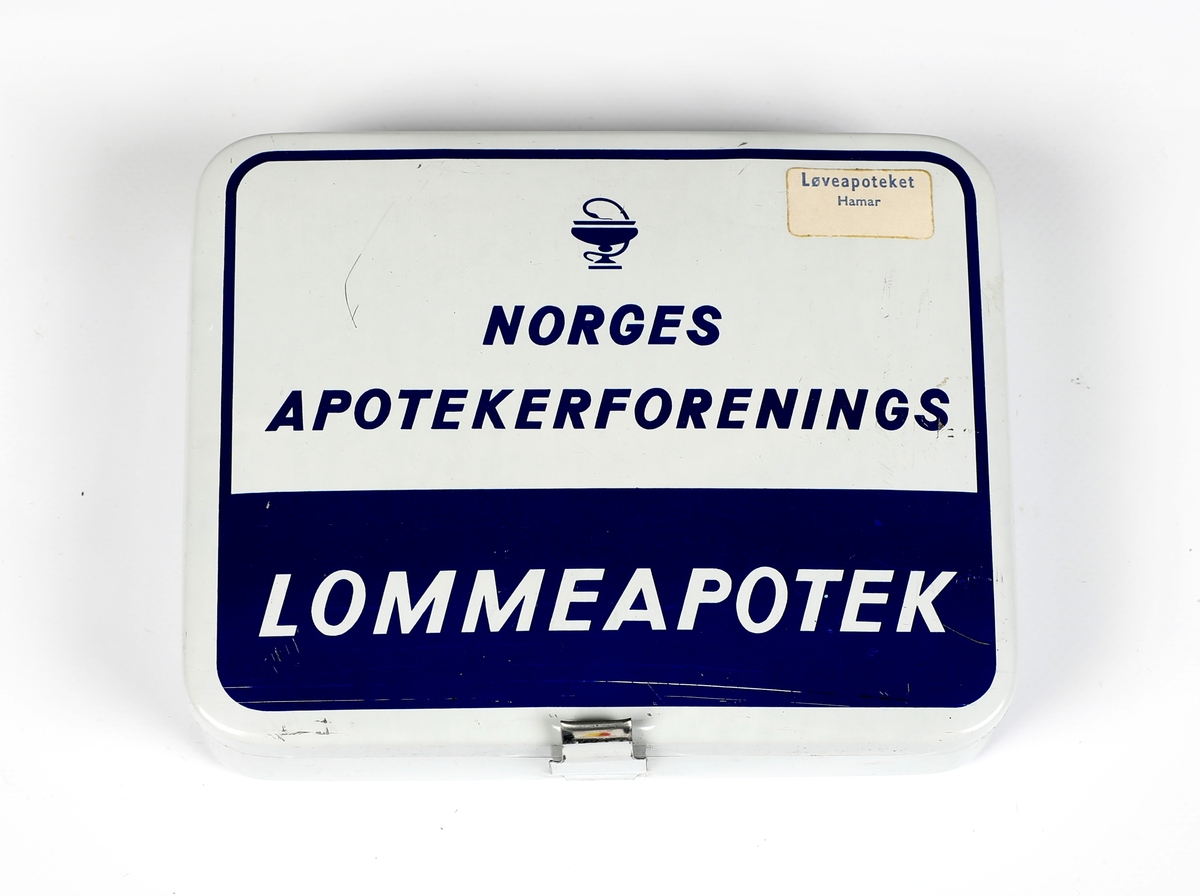 Eske av aluminium med diverse medisinske stoffer, såkalt "lommeapotek". Hvit med blått trykk og klistremerke fra Løveapoteket Hamar. 