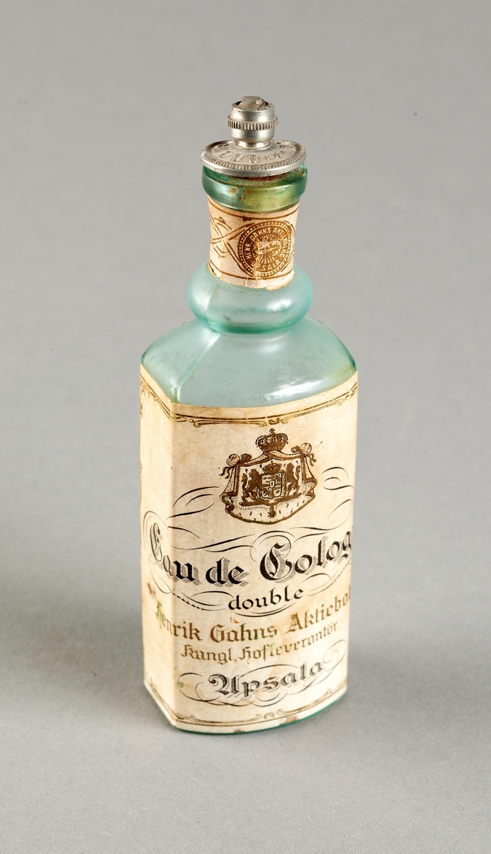 Flaska av svagt grönfärgat glas för eau de cologne, oregelbundet sexkantig med konvex framsida. Tillsluten med kork som är fäst i ett lock av gråaktig metall med text i relief: Gahns Uppsala. Påklistrade pappersetiketter med tryckt text i svart och guld: Eau de Cologne double, Henrik Gahns Aktiebolag, Kungl. Hofleverantör, Upsala. Trasig kork.
