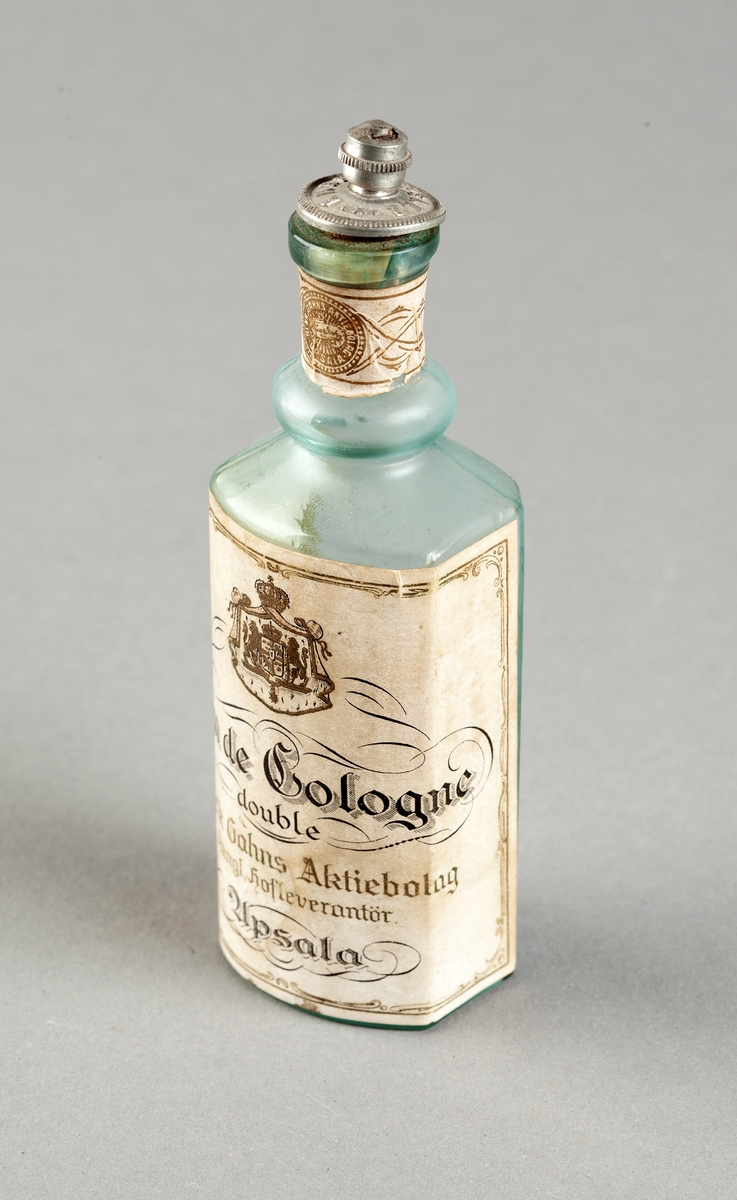 Flaska av svagt grönfärgat glas för eau de cologne, oregelbundet sexkantig med konvex framsida. Tillsluten med kork som är fäst i ett lock av gråaktig metall med text i relief: Gahns Uppsala. Påklistrade pappersetiketter med tryckt text i svart och guld: Eau de Cologne double, Henrik Gahns Aktiebolag, Kungl. Hofleverantör, Upsala. Trasig kork.