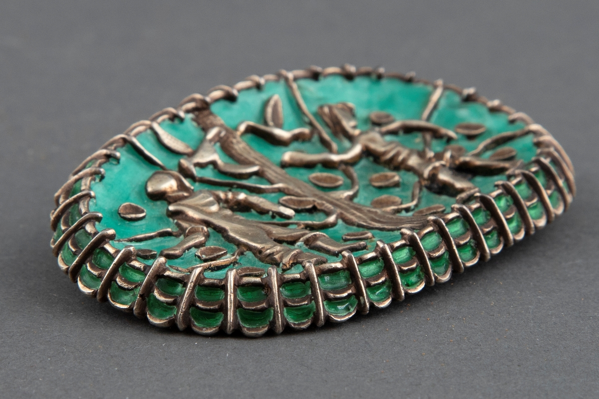 Oval brosje i sølv med turkis emalje. Motiv med to stiliserte menneskefigurer, to fugler og et tre med blader. Nål på baksiden.