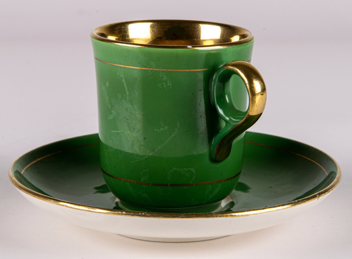 Moccakopp med fat av flintgods, tillverkad av Gefle Porslinsfabrik, modell -AD. Formgivare okänd. Nästan cylindrisk med cirkelformad lodgrepe. Guldfärgad inuti. Enkel grönfärgad dekor utvändigt med två smala guldränder.