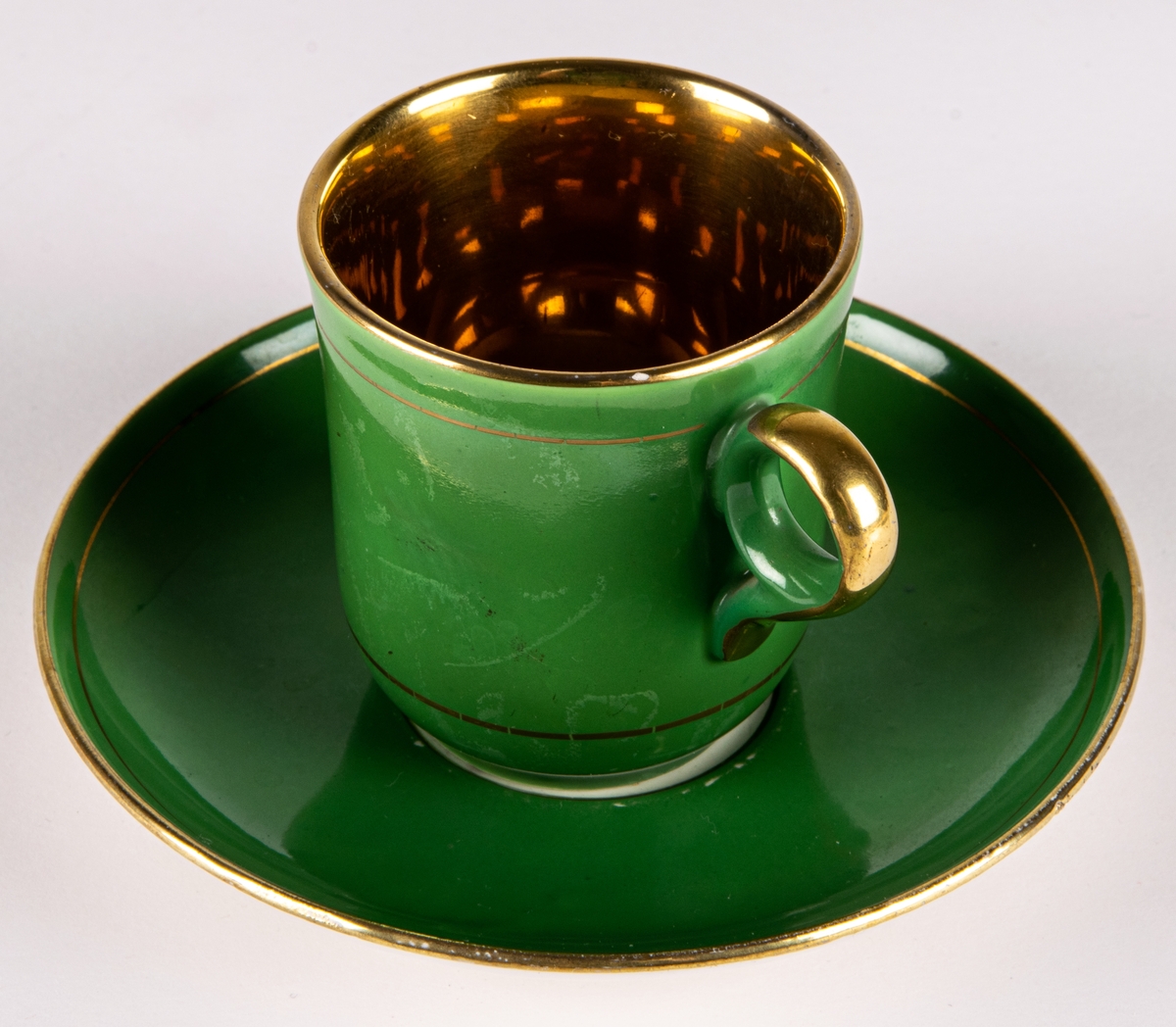 Moccakopp med fat av flintgods, tillverkad av Gefle Porslinsfabrik, modell -AD. Formgivare okänd. Nästan cylindrisk med cirkelformad lodgrepe. Guldfärgad inuti. Enkel grönfärgad dekor utvändigt med två smala guldränder.