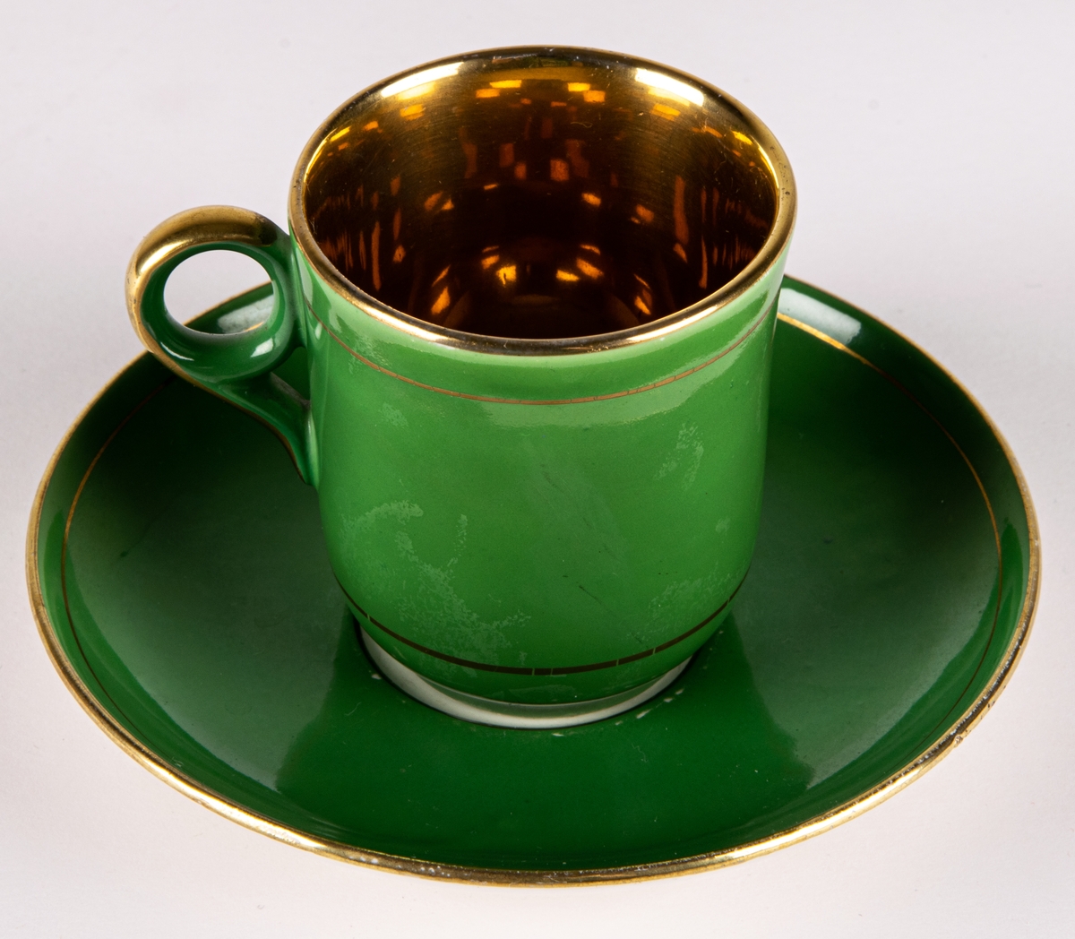 Moccakopp med fat av flintgods, tillverkad av Gefle Porslinsfabrik, modell -AD. Formgivare okänd. Nästan cylindrisk med cirkelformad lodgrepe. Guldfärgad inuti. Enkel grönfärgad dekor utvändigt med två smala guldränder.