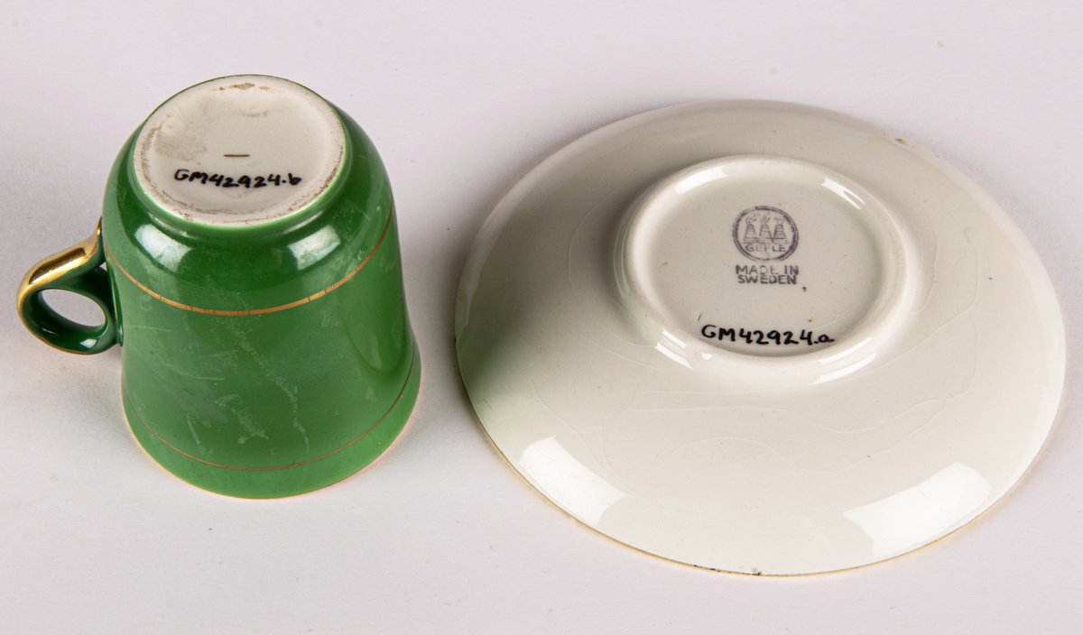 Moccakopp med fat av flintgods, tillverkad av Gefle Porslinsfabrik, modell -AD. Formgivare okänd. Nästan cylindrisk med cirkelformad lodgrepe. Guldfärgad inuti. Enkel grönfärgad dekor utvändigt med två smala guldränder.