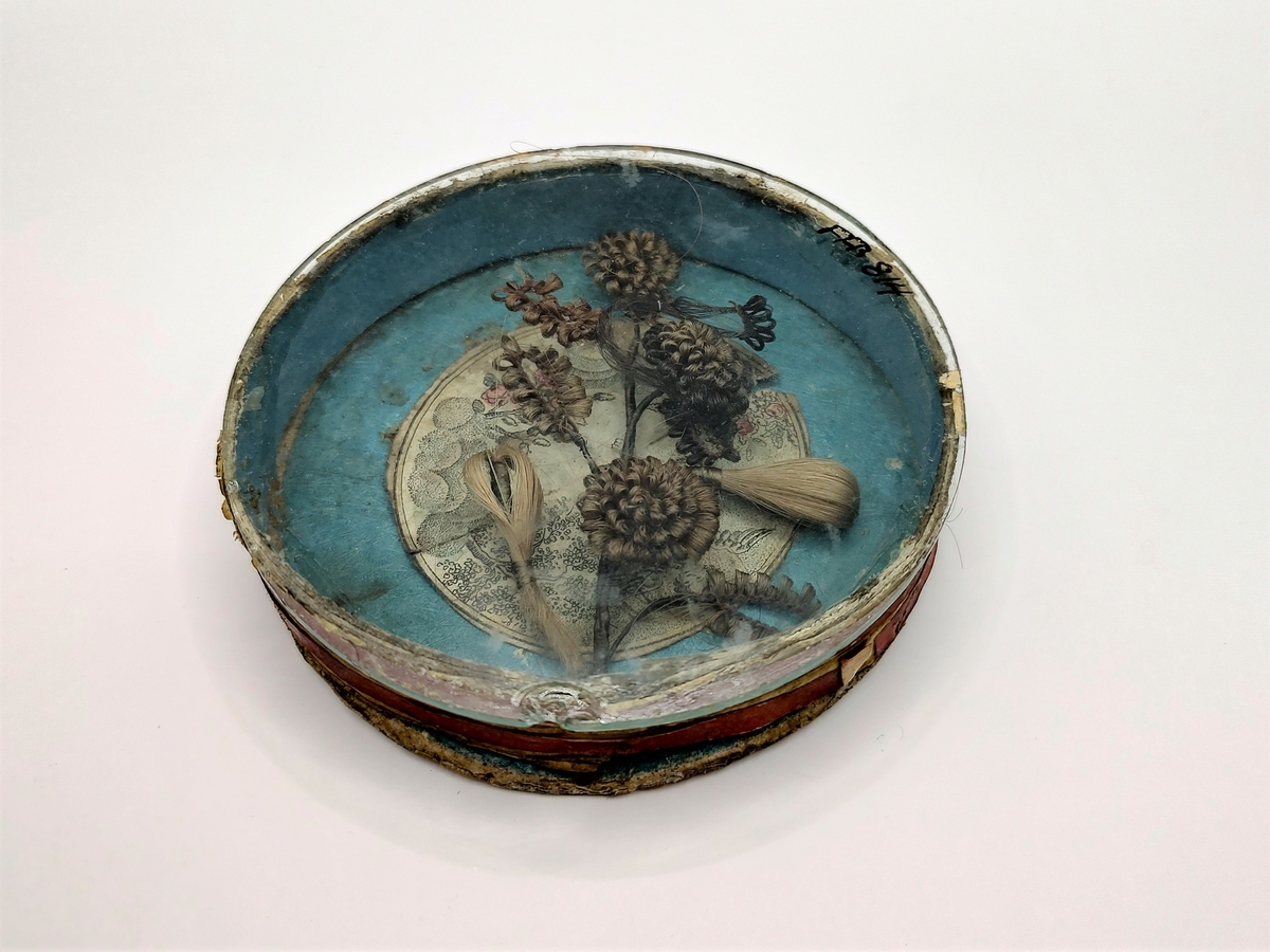 Gren med blomster og blader, laget av flettet hår. Hårarbeidet er plassert inni en sirkulær eske av papp med pålimt glasslokk. Innvendig er esken blåmalt med et pålimt bilde med engel, blomster og skyer i midten. Utvendig er esken rød og hvitstripet. Håret er brunt og blondt.