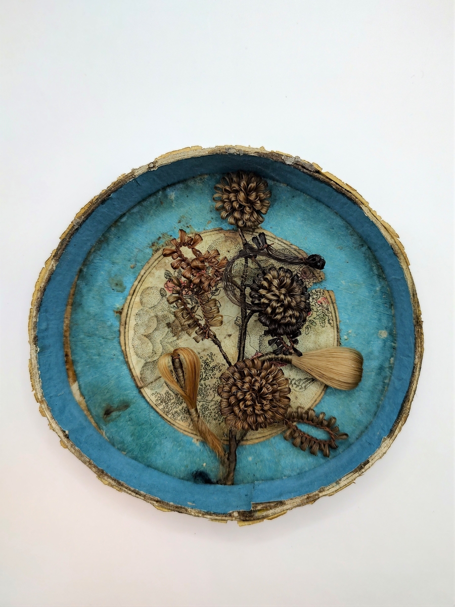 Gren med blomster og blader, laget av flettet hår. Hårarbeidet er plassert inni en sirkulær eske av papp med pålimt glasslokk. Innvendig er esken blåmalt med et pålimt bilde med engel, blomster og skyer i midten. Utvendig er esken rød og hvitstripet. Håret er brunt og blondt.