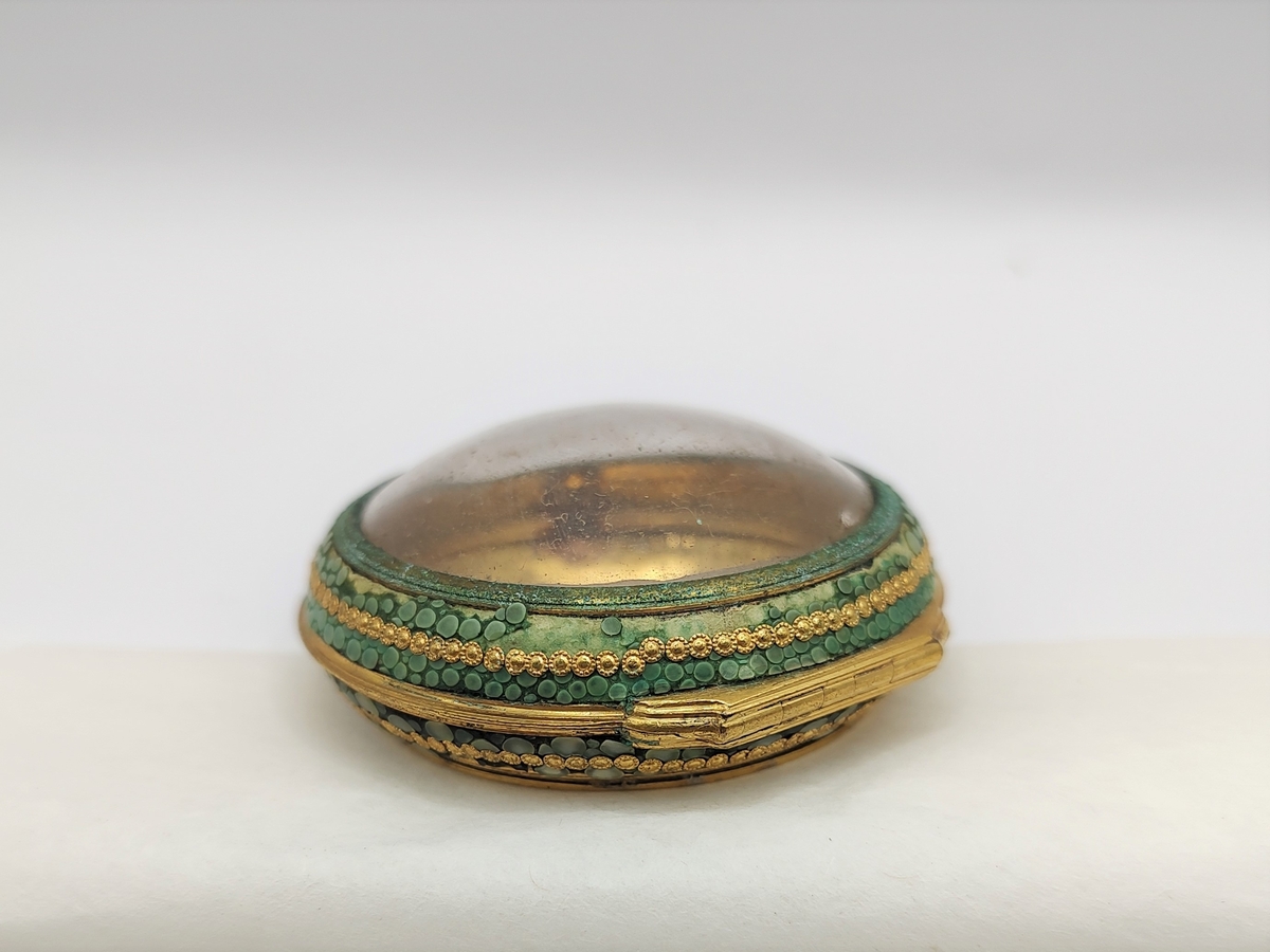 Dåse til lommeur. Sirkulær urkasse av forgylt metall, hengslet lokk med sirkulært glass i midten. Langs kanten er det pålimt grønn mosaikk med forgylt perlekant i midten.