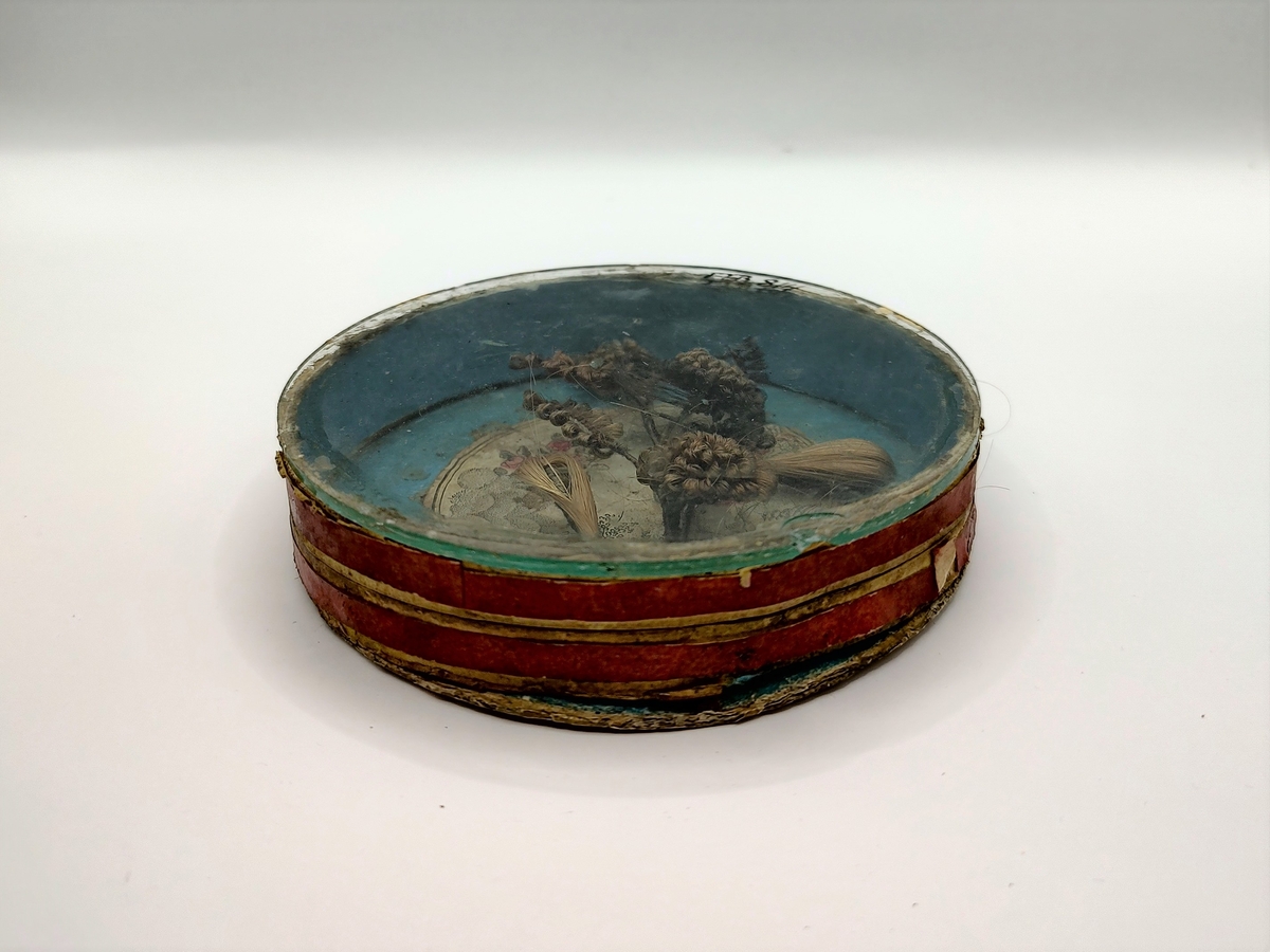 Gren med blomster og blader, laget av flettet hår. Hårarbeidet er plassert inni en sirkulær eske av papp med pålimt glasslokk. Innvendig er esken blåmalt med et pålimt bilde med engel, blomster og skyer i midten. Utvendig er esken rød og hvitstripet. Håret er brunt og blondt.