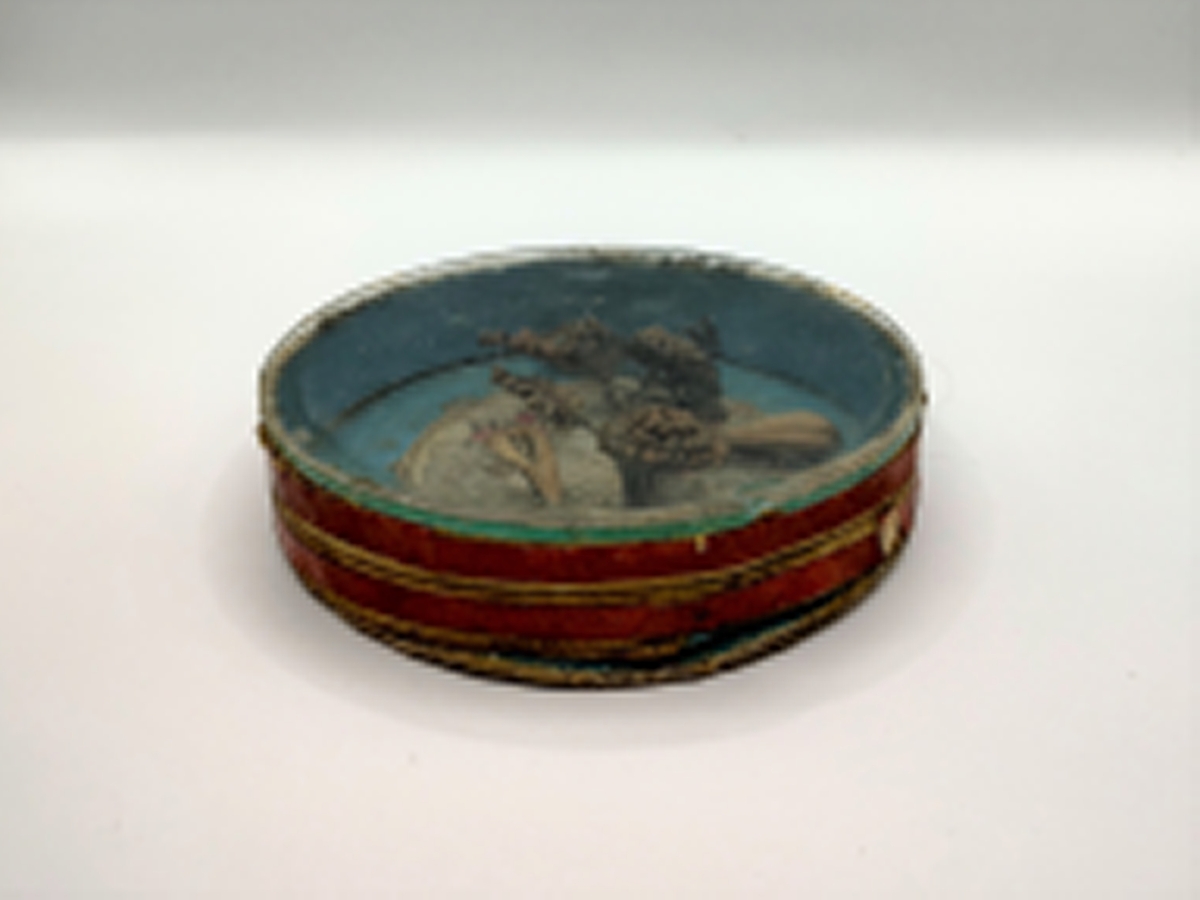 Gren med blomster og blader, laget av flettet hår. Hårarbeidet er plassert inni en sirkulær eske av papp med pålimt glasslokk. Innvendig er esken blåmalt med et pålimt bilde med engel, blomster og skyer i midten. Utvendig er esken rød og hvitstripet. Håret er brunt og blondt.