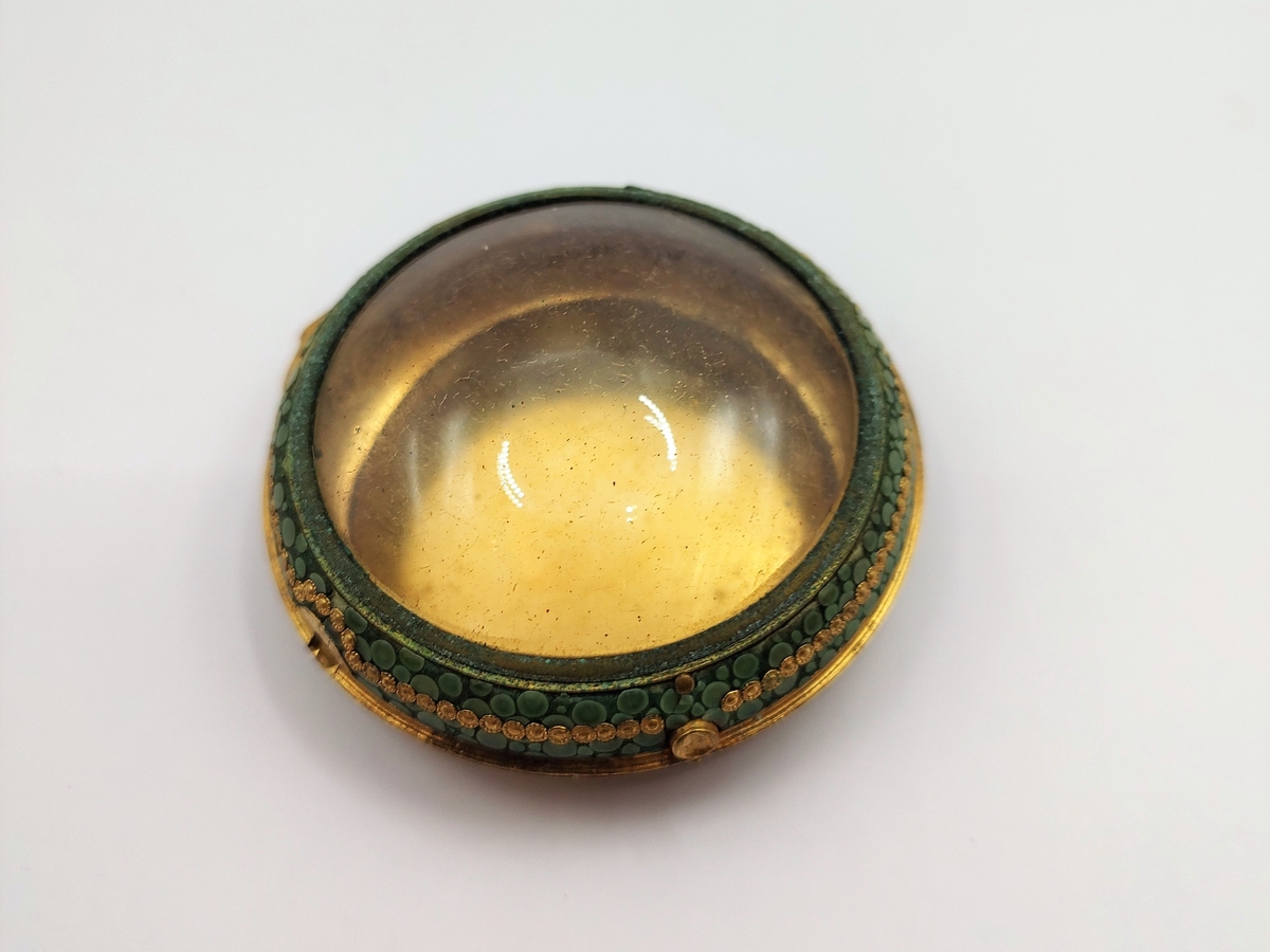 Dåse til lommeur. Sirkulær urkasse av forgylt metall, hengslet lokk med sirkulært glass i midten. Langs kanten er det pålimt grønn mosaikk med forgylt perlekant i midten.