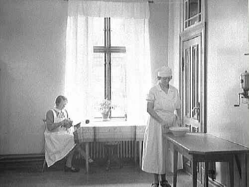 Tjänstefolk hos borgmästare Johan H L Janssen i köksregionen. En kvinna sitter vid fönstret och ser ut att putsa en kopparkittel medan den andra står med en bunke vid ett bord. På väggen sitter en kaffekvarn. Våningen låg i "tvillinghuset" i kv Komedianten 1. Janssen flyttade dit 1904 med familjen och förvärvade fastigheten. (Hustrun dog 1940 så troligen är bilden tagen dessförinnan.)