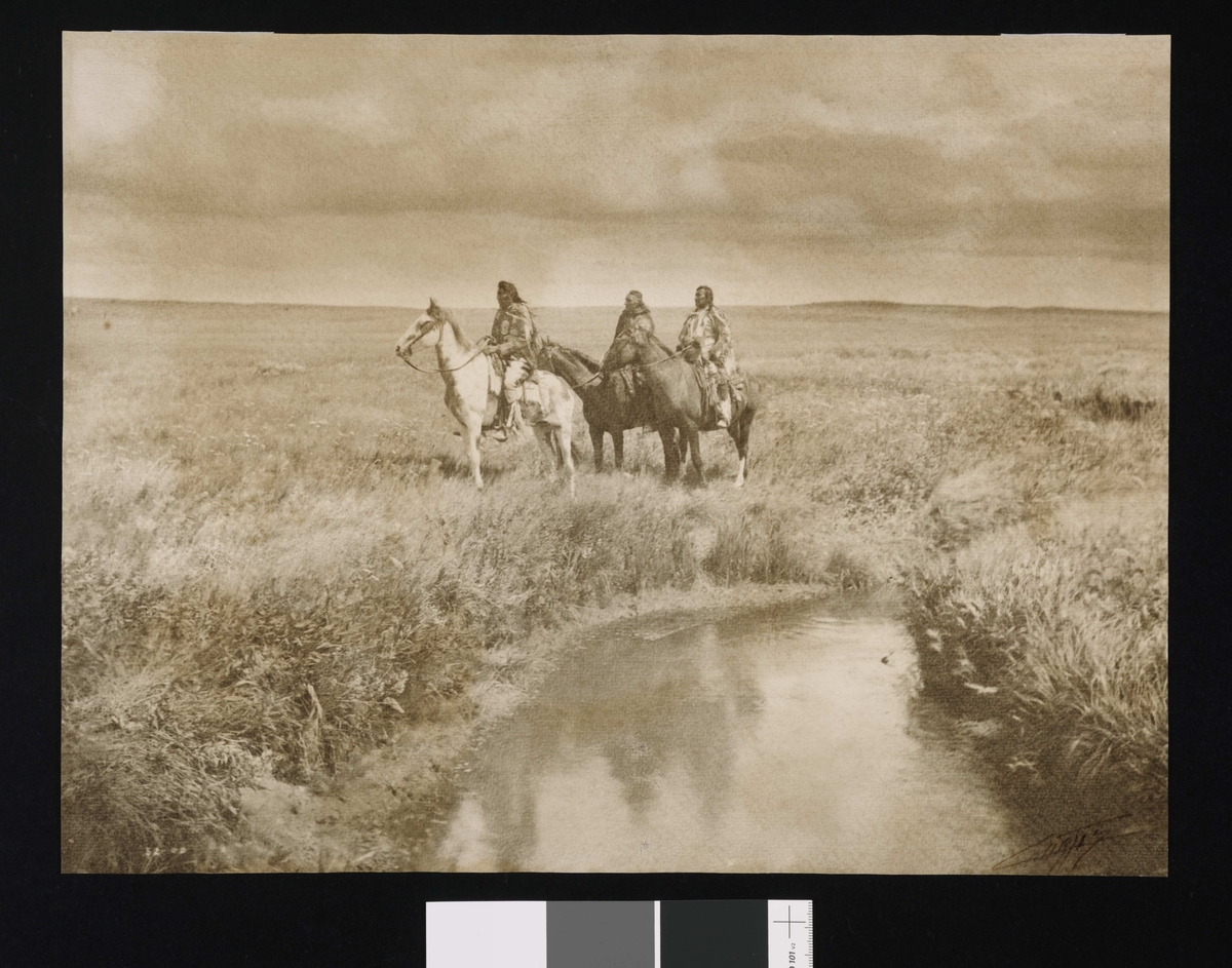 Tre menn til hest, prærielandskap. Blackfoot-høvdinger kledd i tradisjonelt antrekk.