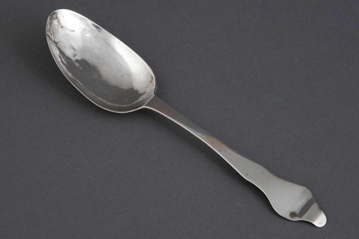 Spiseskje med stort, ovalt skjeblad og fiolinskaft med tungeformet opp- og bakoverbøyd ende. Enkel strekdekor i rokokkostil på skjebladets underside.