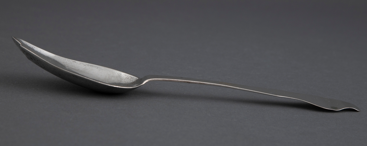 Spiseskje med stort, ovalt skjeblad og fiolinskaft med tungeformet opp- og bakoverbøyd ende. Enkel strekdekor i rokokkostil på skjebladets underside.