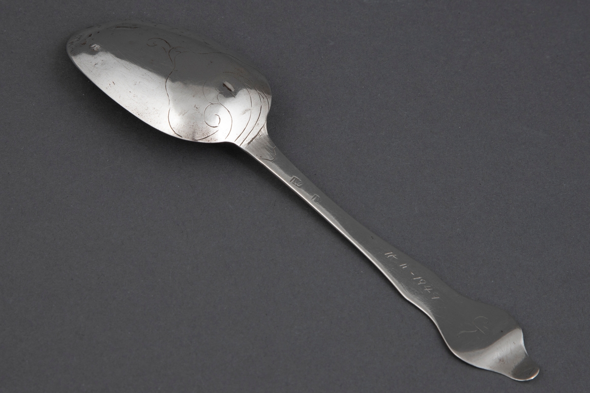 Spiseskje med stort, ovalt skjeblad og fiolinskaft med tungeformet opp- og bakoverbøyd ende. Enkel strekdekor i rokokkostil på skjebladets underside.