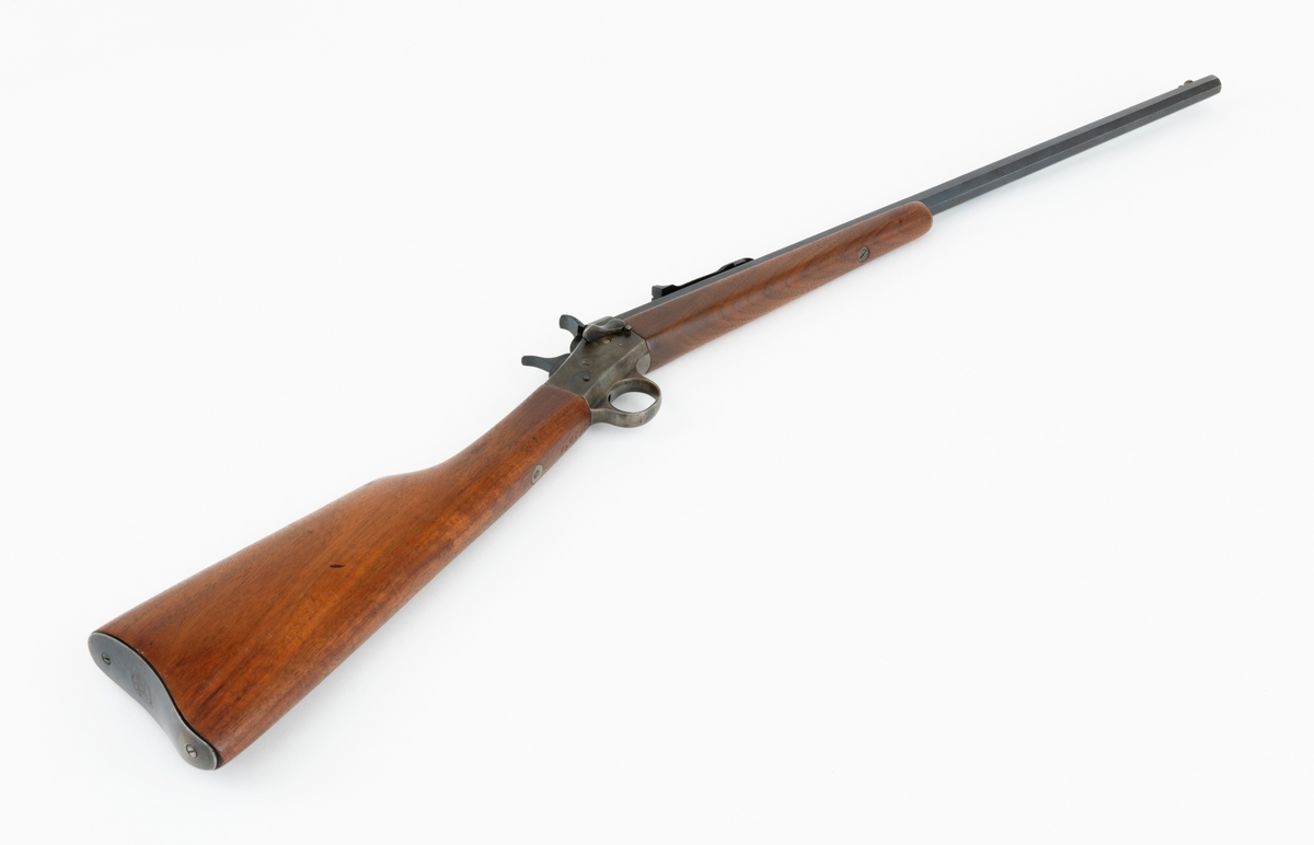 Tillegg innskrift: Remington TRADE MARK MODEL 4 25-10 R. F. Geværet har brunt, oljebehandlet treverk. Framstillingsnummeret er J 304870. Laget ca. 1870. Av størrelse kan geværet se ut som ei salongrifle, men kaliberet, 25-10, er litt større enn på Krag. R. F. står for Rand Fire (randtenning. ) 10-tallet står for 10 gram svartkrutt. Dvs. ca. 4 gram. Dette er en meget kraftig ladning, nesten like mye som på den store Remington. Det må være en av de raskeste svartkruttpatroner som er laget.