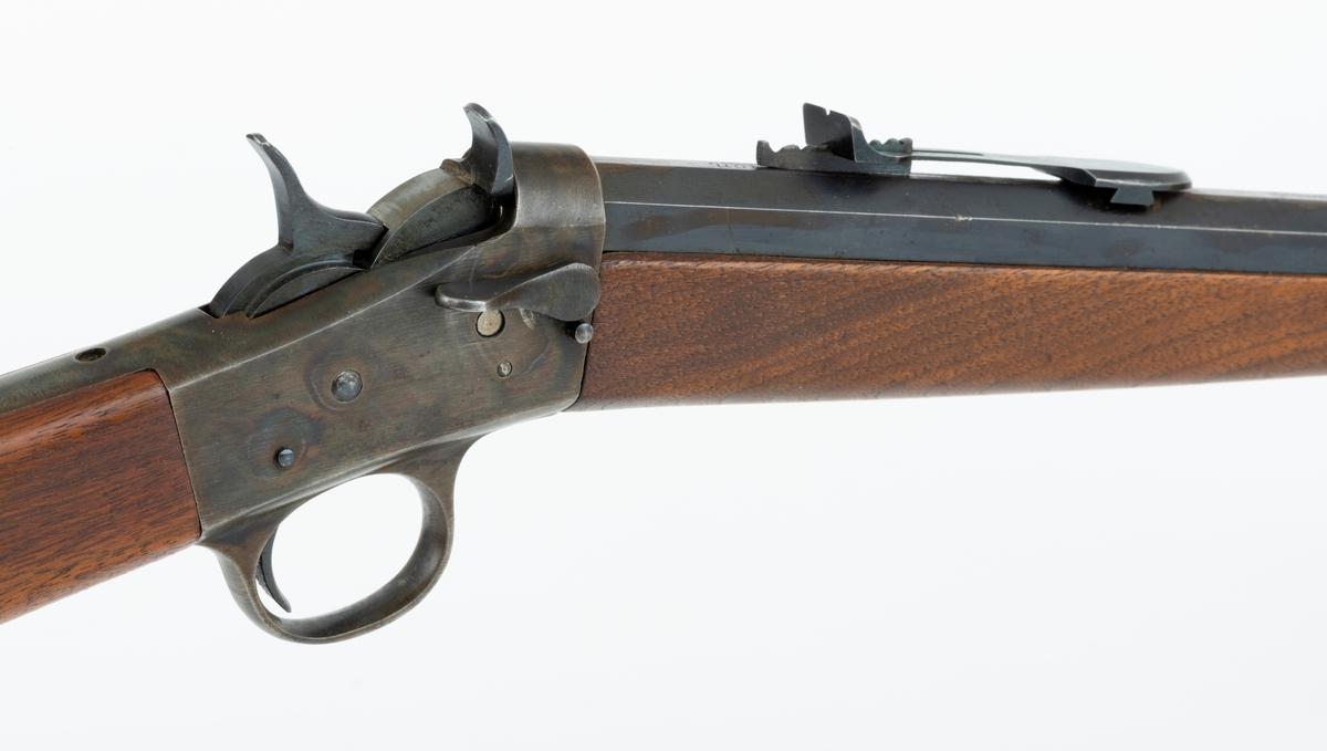 Tillegg innskrift: Remington TRADE MARK MODEL 4 25-10 R. F. Geværet har brunt, oljebehandlet treverk. Framstillingsnummeret er J 304870. Laget ca. 1870. Av størrelse kan geværet se ut som ei salongrifle, men kaliberet, 25-10, er litt større enn på Krag. R. F. står for Rand Fire (randtenning. ) 10-tallet står for 10 gram svartkrutt. Dvs. ca. 4 gram. Dette er en meget kraftig ladning, nesten like mye som på den store Remington. Det må være en av de raskeste svartkruttpatroner som er laget.